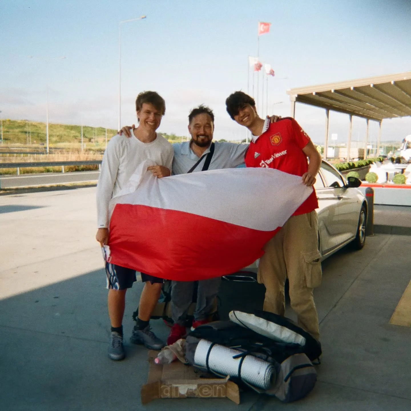 Three people holding a Polish flag next to a car, backpacks at their feet